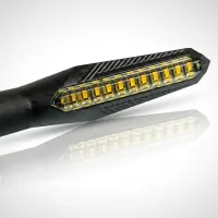 gros plan sur un clignotant LED séquentiel Next‑Tech pour moto, optique orange et boîtier noir sur fond studio blanc dégradé gris.
