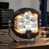 Phare LED rond additionnel Next‑Tech allumé avec feu de position orange, posé sur un établi de garage en vue de face